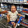 baby, child, shopping_cart, smile, person, store, aisle, bottles, cleaning_supplies, hand, white_clothing, blue_shorts, footwear, woven_bag, hair_tie, happy, indoor, retail, metal_cart, curly_hair