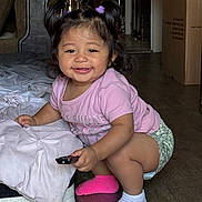 Rosie is registered to the contest to win money with this photo: baby, cardboard_box, child, cushion, cute, floor, furniture, hair_ties, happy, indoor, mismatched_socks, patterned_shorts, person, pigtails, pink_shirt, playful, remote_control, smiling, toddler, wooden_floor