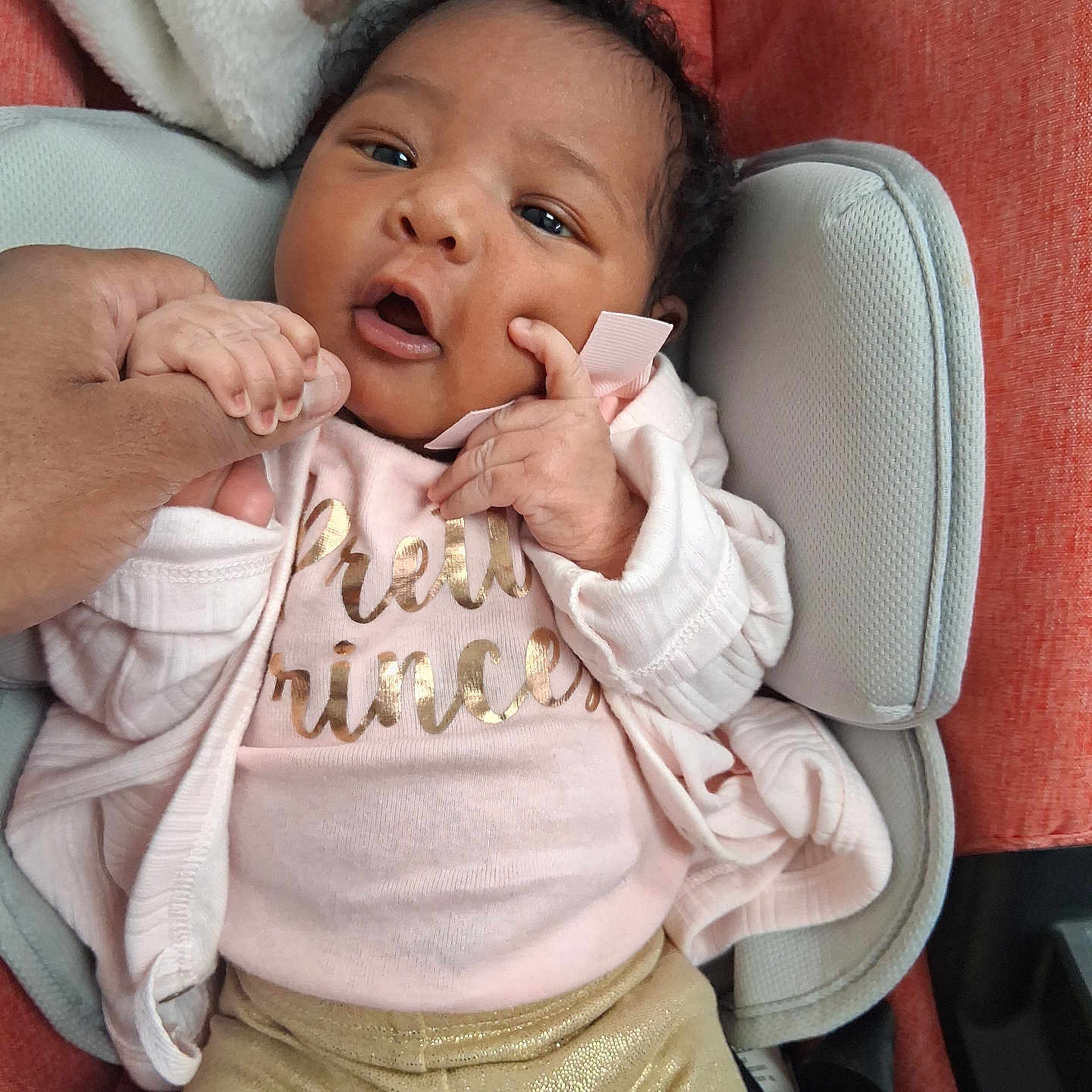 Kyra'Yana joined the competition — help win amazing prizes! baby, infant, child, portrait, sitting, car_seat, pink_clothing, sparkly_pants, blanket, cushion, adult_hand, finger, open_mouth, wide_eyes, curly_hair, indoor, close_up, cozy, text_on_clothing, happy