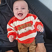 Samaël participe au concours pour gagner de l'argent avec cette photo : adult_hand, baby, casual_clothing, child, couch, happy, home, human, indoor, infant, joyful, orange, pants, person, portrait, seated, smiling, socks, striped_clothing, sweater