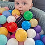 baby, child, ball_pit, colorful_balls, indoor, curious, wide_eyes, clothing, person, hand, face, head, play, toy, seated, soft_lighting, background, adult, cushion, cozy