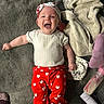 baby, infant, smiling, red_pants, heart_pattern, headband, lying_down, carpet, blanket, baby_bottle, happy, cute, child, toddler, clothing, indoor, person, cozy, footwear, soft_toys
