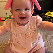 Sofia is registered to the contest to win money with this photo: arms_outstretched, baby, bunny_ears, child, cute, dress, floor, furniture, happy, headband, indoor, infant, patterned_surface, person, pink, playful, room, sitting, smiling, toy