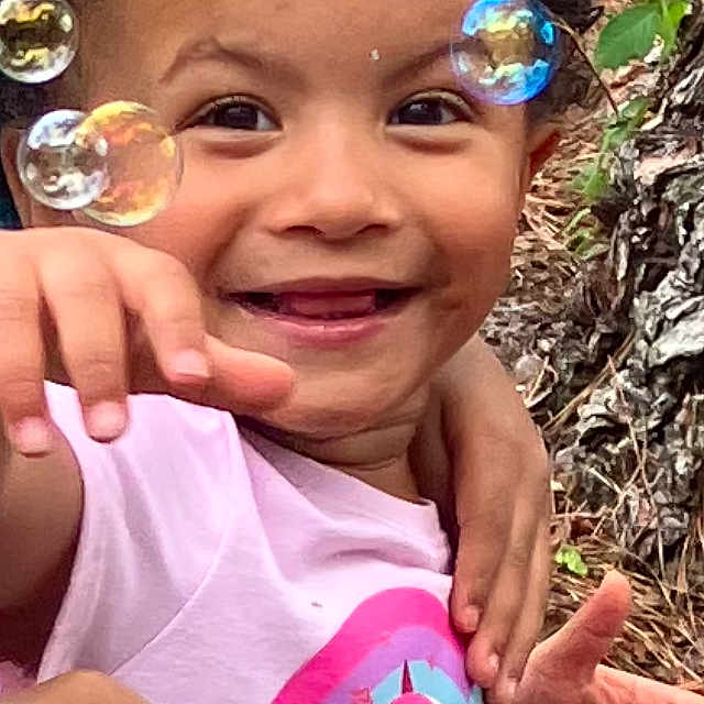 Zaria is registered to the contest to win money with this photo: bubbles, cartoon_sharks, child, closeup, curly_hair, expression, face, hand, happy, joyful, leaves, nature, outdoor, person, pink_shirt, playing, pouch, smiling, sunlight, tree_bark
