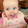 Greta is registered to the contest to win money with this photo: baby, child, high_chair, bib, pink_clothing, rainbow, hearts, tongue_out, happy, cute, indoor, restaurant, booth, wooden_furniture, playful, funny_hair, face, smiling, person, infant