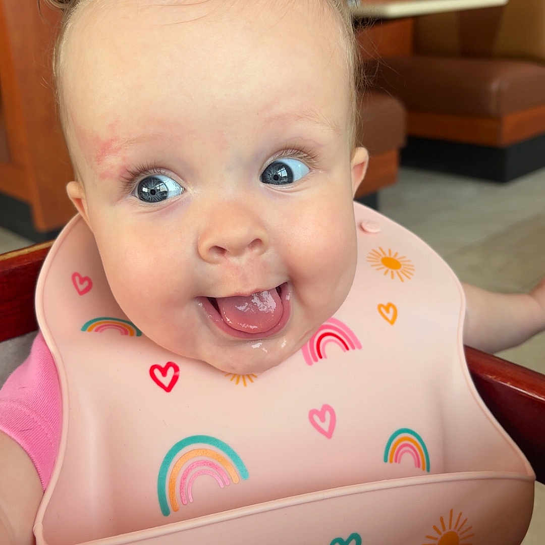 Greta is registered to the contest to win money with this photo: baby, child, high_chair, bib, pink_clothing, rainbow, hearts, tongue_out, happy, cute, indoor, restaurant, booth, wooden_furniture, playful, funny_hair, face, smiling, person, infant