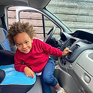 Karel participe au concours pour gagner de l'argent avec cette photo : child, curly_hair, red_hoodie, blue_pants, sneakers, car_interior, dashboard, car_seat, car_door, window, steering_wheel, smiling, playful, young_child, indoor, wooden_fence, casual_clothing, seat_cushion, footwear, person
