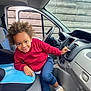 child, curly_hair, red_hoodie, blue_pants, sneakers, car_interior, dashboard, car_seat, car_door, window, steering_wheel, smiling, playful, young_child, indoor, wooden_fence, casual_clothing, seat_cushion, footwear, person