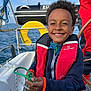 child, smile, life_jacket, rope, boat, sailboat, ocean, sky, water, yellow, blue, red, person, nautical, outdoor, happy, youth, curly_hair, clothing, marine