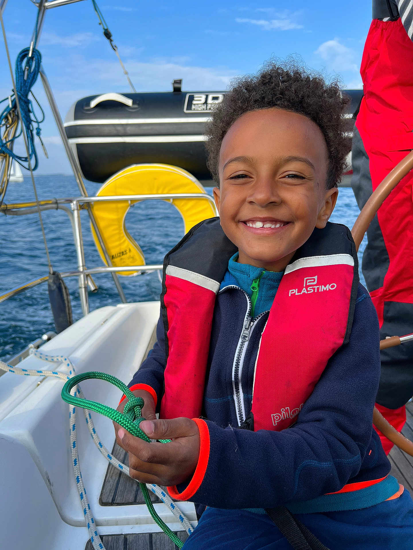 Owen participe au concours pour gagner de l'argent avec cette photo : child, smile, life_jacket, rope, boat, sailboat, ocean, sky, water, yellow, blue, red, person, nautical, outdoor, happy, youth, curly_hair, clothing, marine