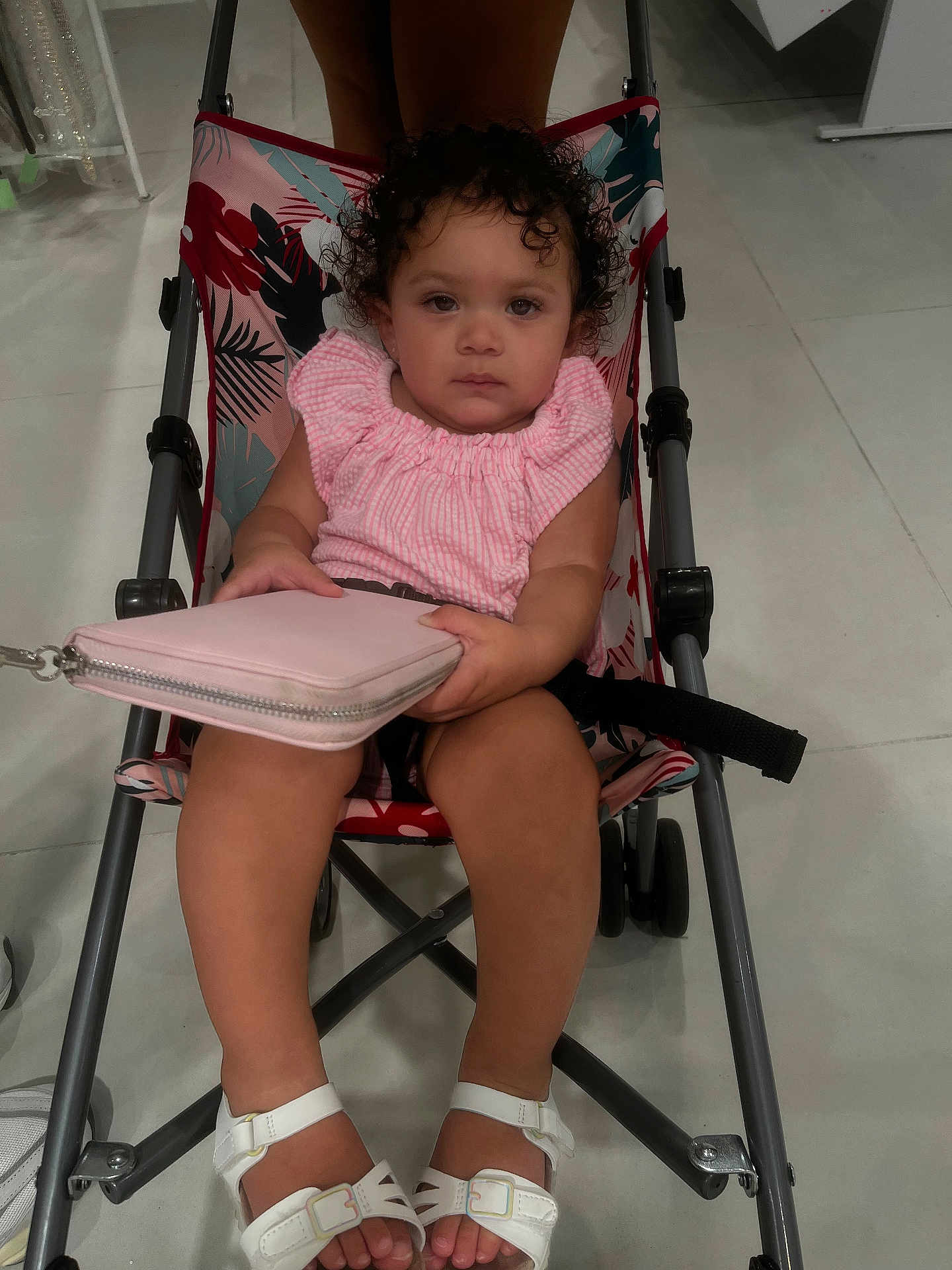 Olivianna joined the competition — help win amazing prizes! toddler, child, stroller, pink_clothing, curly_hair, white_sandals, wallet, indoor, floor, tile_floor, baby, person, sitting, serious_expression, footwear, seat, baby_stroller, handheld_object, clothing, young_child