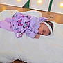 baby, infant, sleeping, headband, purple_clothing, butterfly_pattern, white_blanket, wooden_surface, decorative_lights, indoor, cozy, newborn, peaceful, portrait, lying_down, soft_texture, sleeping_pose, resting, tiny_hand, newborn_outfit