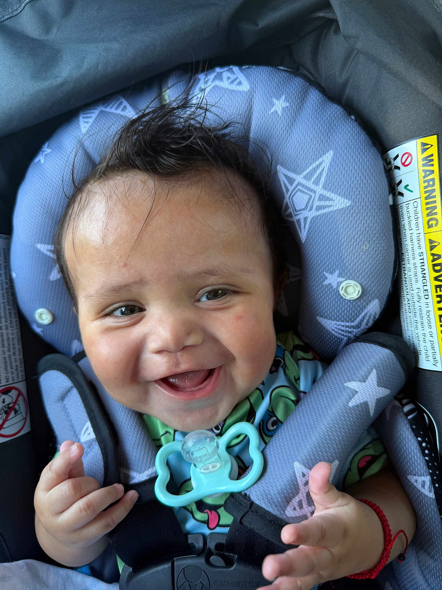 Donovan is registered to the contest to win money with this photo: baby, smiling, car_seat, pacifier, star_pattern, child, happy, infant, clothing, seatbelt, face, hand, cute, young_child, safety, indoors, person, closeup, expression, red_bracelet