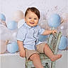 toddler, child, smile, blue_eyes, checkered_shirt, beige_shorts, wooden_chair, pastel_colors, balloons, birthday_theme, studio_photo, indoor, cute, happy, baby, portrait, sitting, playful, light_background, rustic_furniture