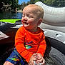 blue_sky, child, clothing, colorful_shorts, daylight, happy, holding_object, inflatable_tube, nature, orange_shirt, outdoor, playing, river, sitting, smiling, summer, sunlight, toddler, trees, water