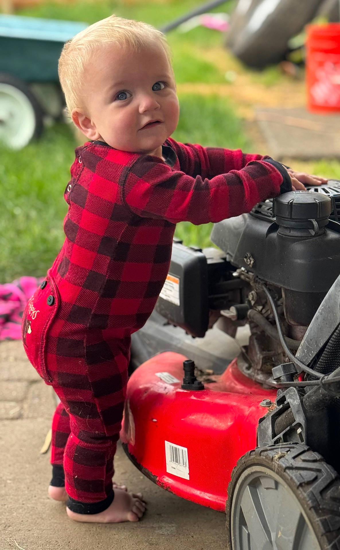 baby, barefoot, checkered_clothing, child, concrete, curious, footwear, garden, grass, lawn_mower, machine, outdoor, person, playful, red_onesie, standing, toddler, toy, wheel, young_child