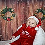 baby, child, santa_hat, red_clothing, christmas_wreath, holiday, festive, smiling, happy, snowflakes, wooden_background, indoor, seasonal, decorations, christmas_tree, fluffy_chair, portrait, cute, celebration, winter