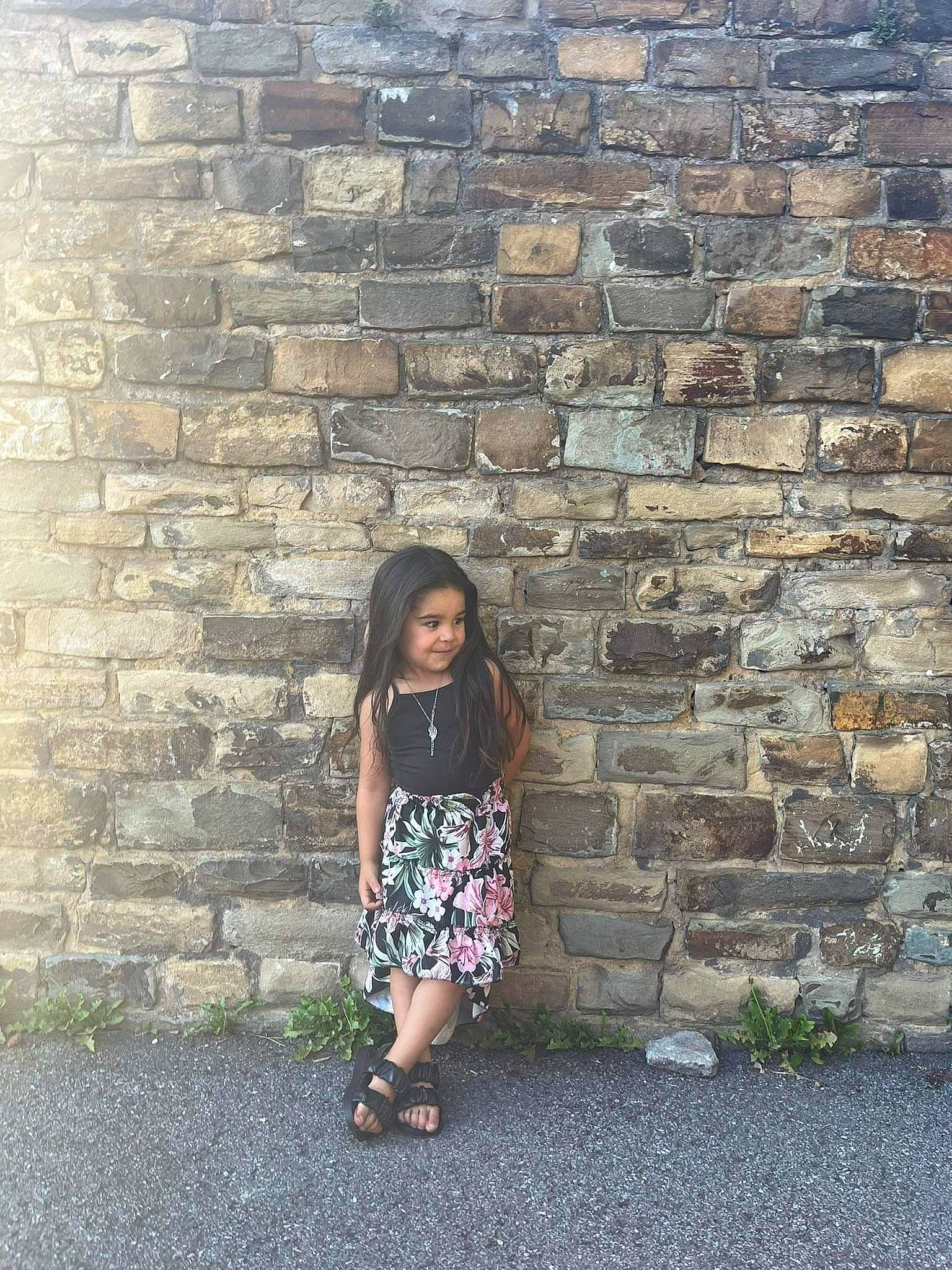 Eliyana participe au concours pour gagner de l'argent avec cette photo : asphalt, brick, brickwork, dress, grass, grey, hair, hairstyle, human, line, pattern, person, plant, public_space, road_surface, sandal, sidewalk, sitting, standing, stone_wall
