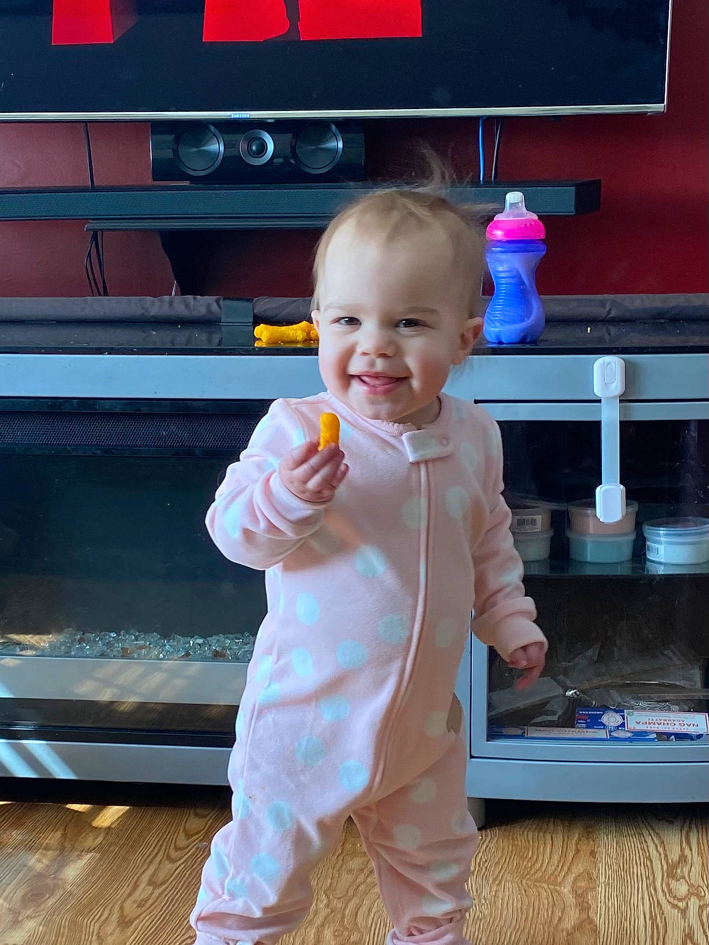 Remmington is registered to the contest to win money with this photo: baby, baby_toddler_clothing, child, finger, flooring, fun, gesture, hand, happy, hardwood, home_appliance, joy, orange, person, skin, sleeve, smile, standing, television, thumb