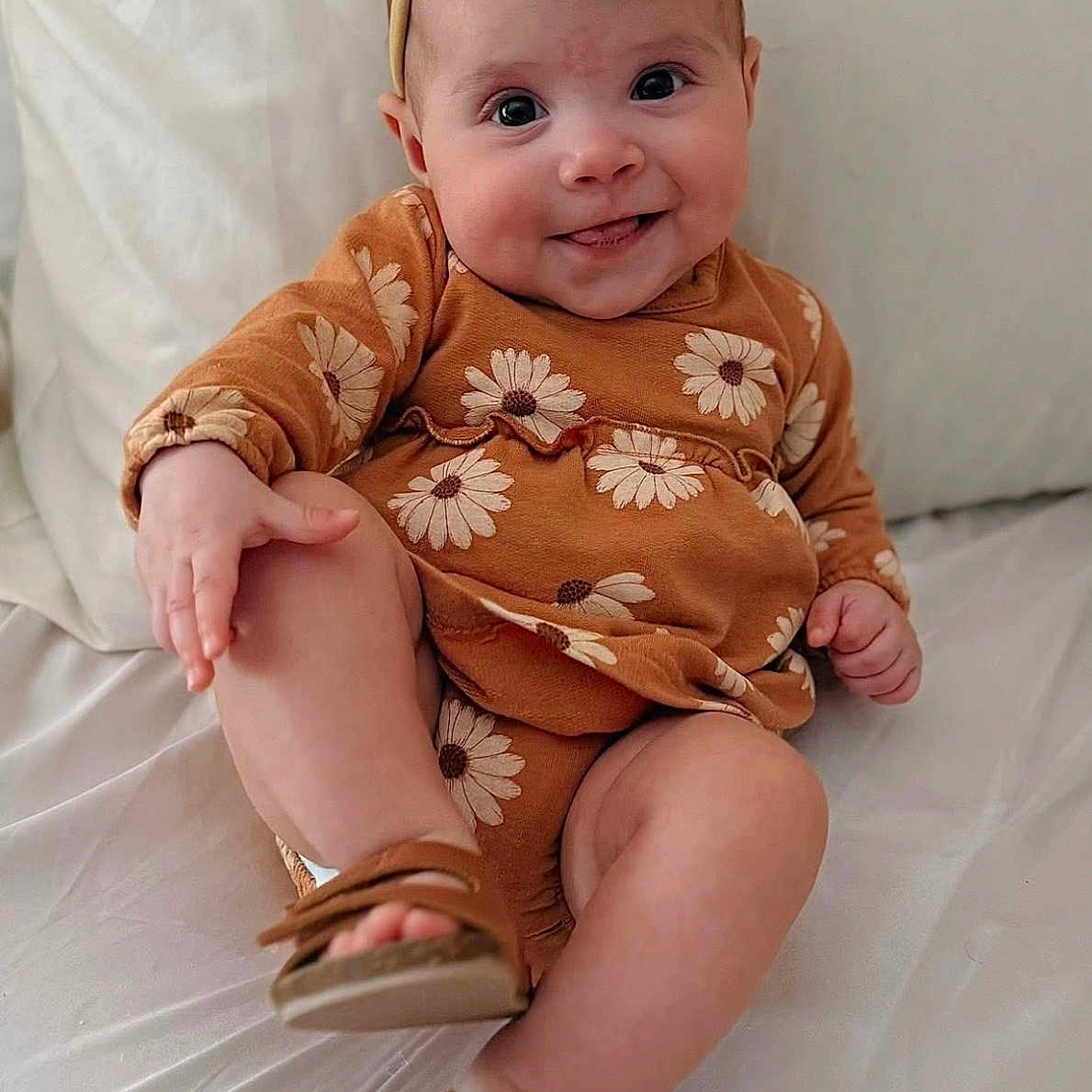 Mila is registered to the contest to win money with this photo: baby, child, infant, smile, headband, flower, daisy, brown_clothing, sandals, sitting, bed, white_bed, cute, happy, portrait, skin, face, hand, footwear, indoors