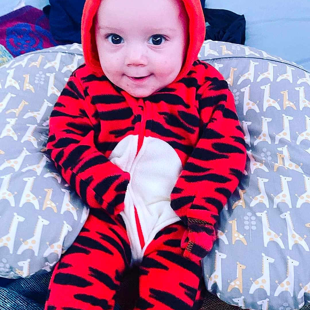 Lucca joined the competition — help win amazing prizes! baby, child, cozy, cute, face, giraffe_pattern, happy, head, hood, indoor, infant, onesie, person, pillow, plush, red_clothing, seated, smile, tiger_stripes, young_child
