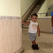 Yassir a rejoint le concours — aidez-le/la à gagner de superbes lots ! background, casual_pose, child, clothing, floor, footwear, handrail, indoor, leaning, person, portrait, shoes, shorts, sleeveless_shirt, smiling, stairs, standing, tile, toddler, wall