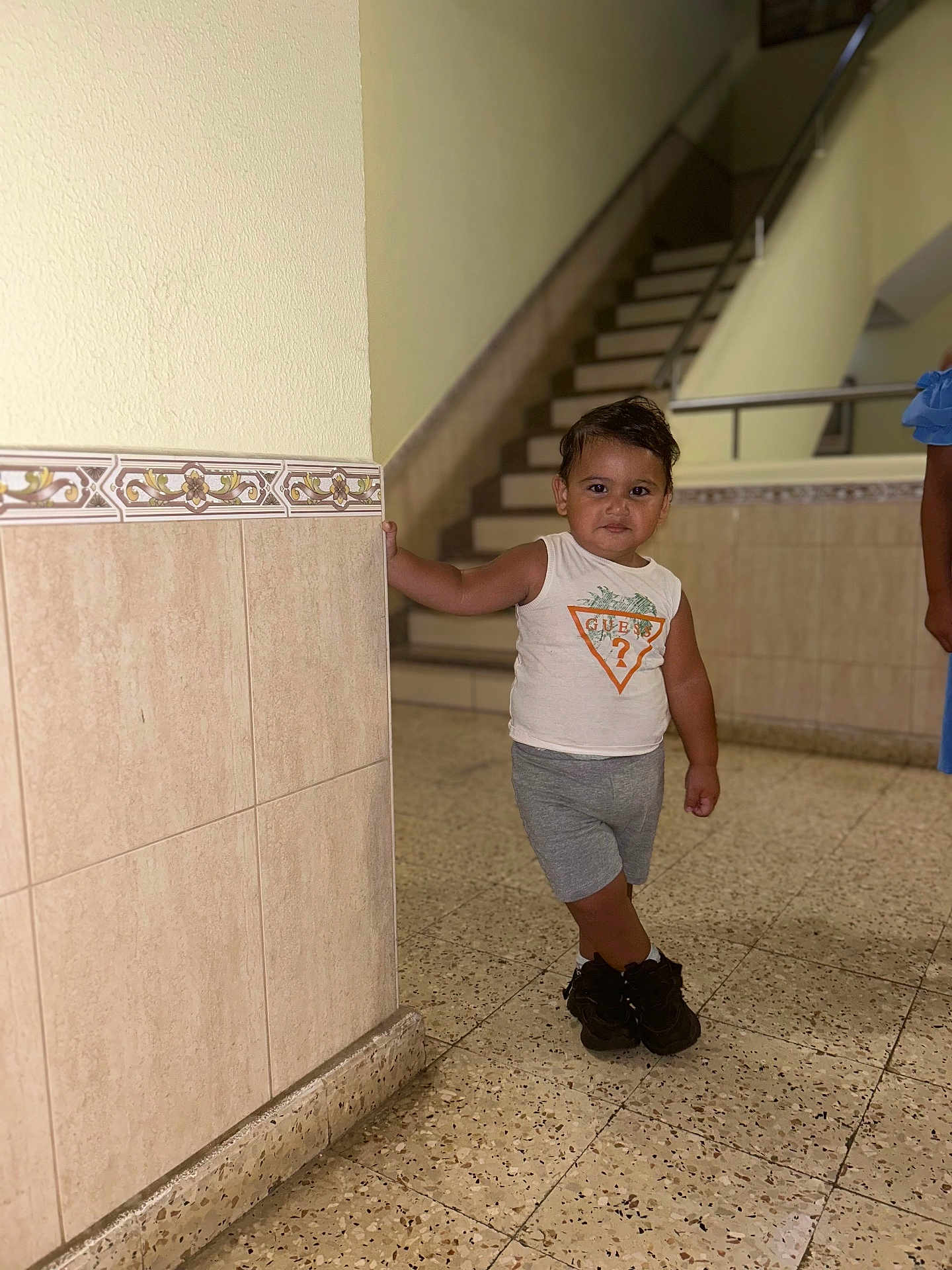 Yassir a rejoint le concours — aidez-le/la à gagner de superbes lots ! background, casual_pose, child, clothing, floor, footwear, handrail, indoor, leaning, person, portrait, shoes, shorts, sleeveless_shirt, smiling, stairs, standing, tile, toddler, wall