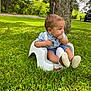 baby, child, curious, daytime, footwear, grass, greenery, hand_in_mouth, lawn, nature, outdoor, park, road_sign, seat, shirt, side_view, sunlight, suspenders, tree, vehicle