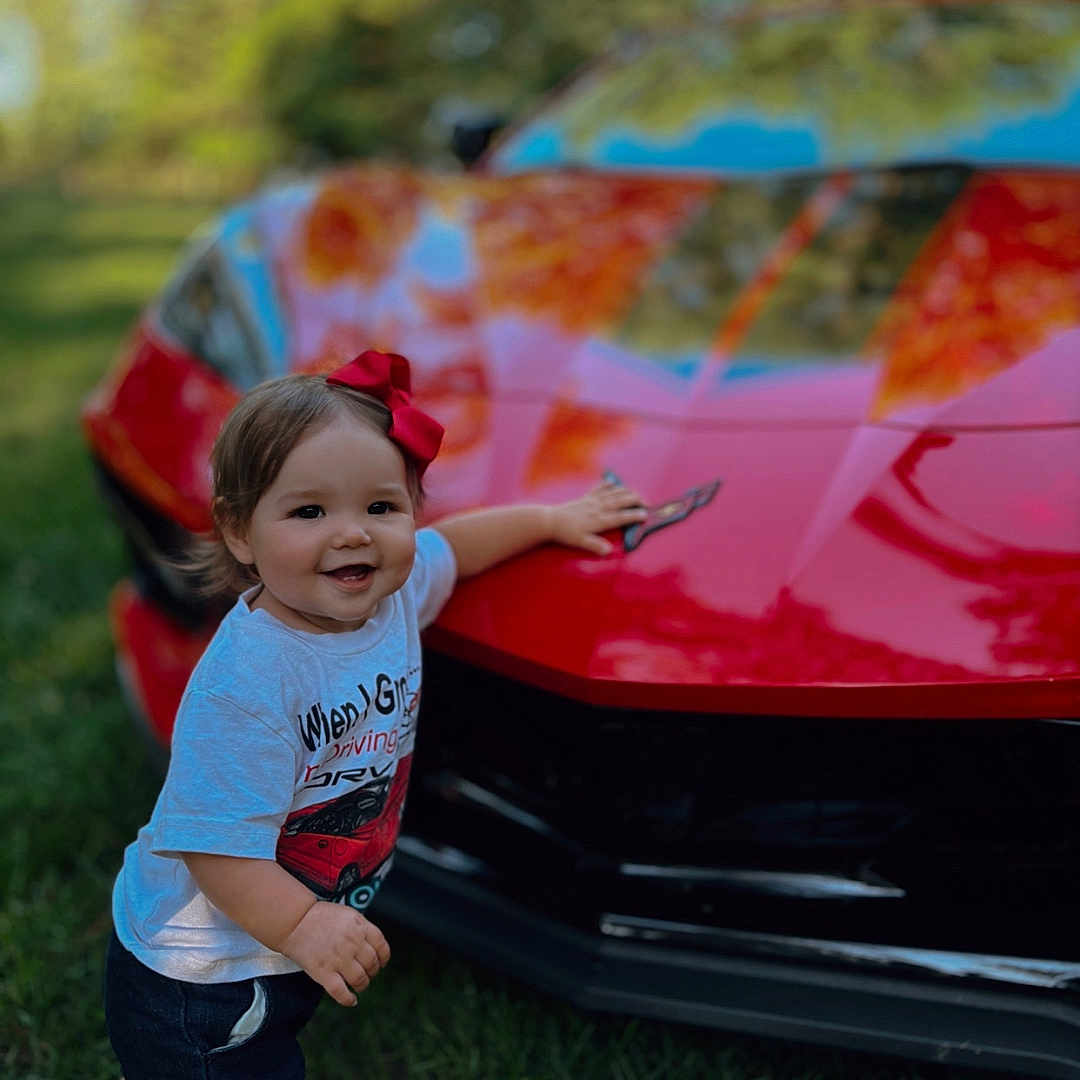 Nyla joined the competition — help win amazing prizes! barefoot, car, child, cute, daylight, front_bumper, grass, happy, nature, outdoor, person, portrait, red_bow, red_car, reflection, shorts, smiling, sports_car, toddler, white_tshirt