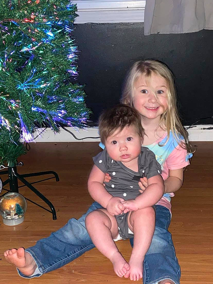 Emberleigh joined the competition — help win amazing prizes! christmas_ornament, christmas_tree, event, flooring, foot, fun, happy, human_body, jeans, joy, lap, leg, leisure, person, sitting, smile, snapshot, standing, thigh, toddler