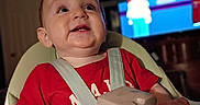 Ian is registered to the contest to win money with this photo: baby, child, high_chair, red_shirt, toys, smiling, happy, indoor, television, cartoon, face, person, seat, plastic_toy, table, room, furniture, infant, expression, entertainment