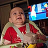 baby, child, high_chair, red_shirt, toys, smiling, happy, indoor, television, cartoon, face, person, seat, plastic_toy, table, room, furniture, infant, expression, entertainment