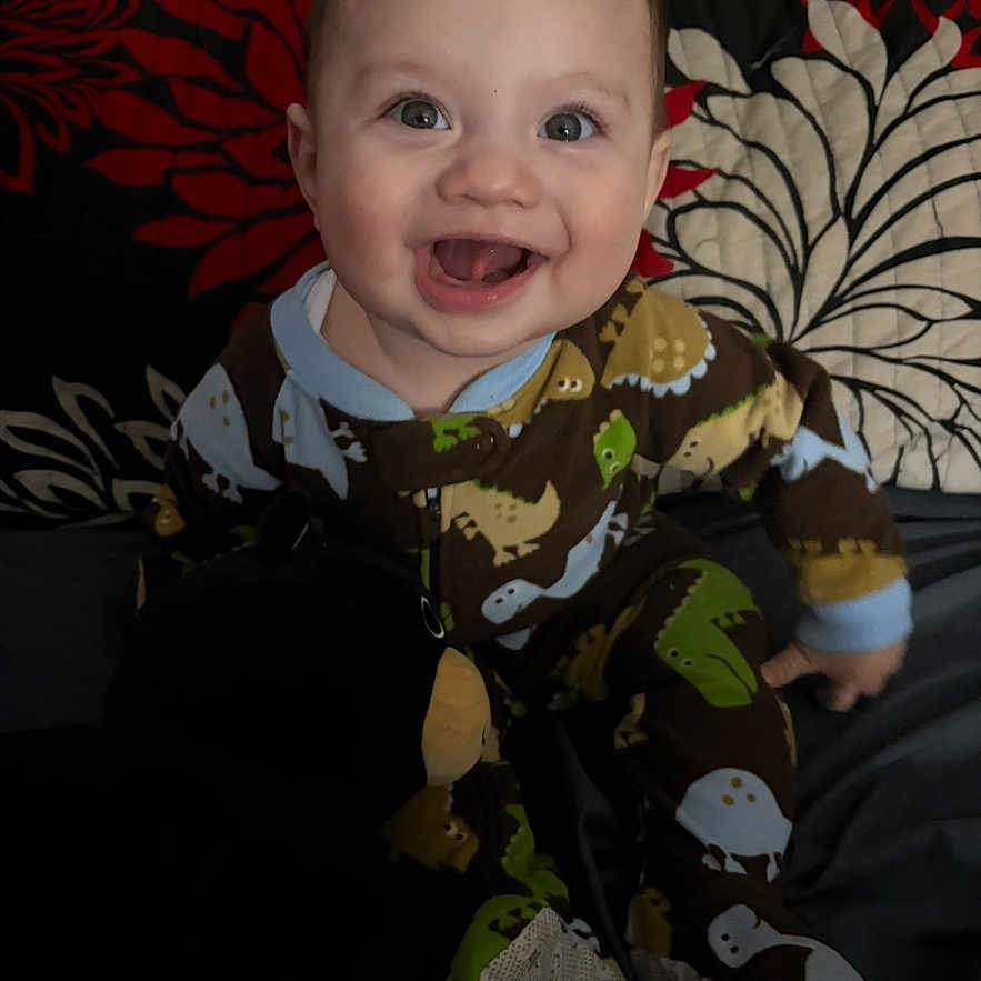 Zaid is registered to the contest to win money with this photo: baby, child, smiling, dinosaur_onesie, sitting, indoor, happy, face, expression, pillow, pattern, floral, cute, infant, portrait, clothing, eyes, mouth_open, cheerful, young