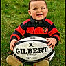 toddler, child, grass, rugby_ball, smile, happy, outdoor, red_sweater, black_sweater, baby, playing, toy, sport, sitting, cute, portrait, person, fun, daylight, casual_clothing