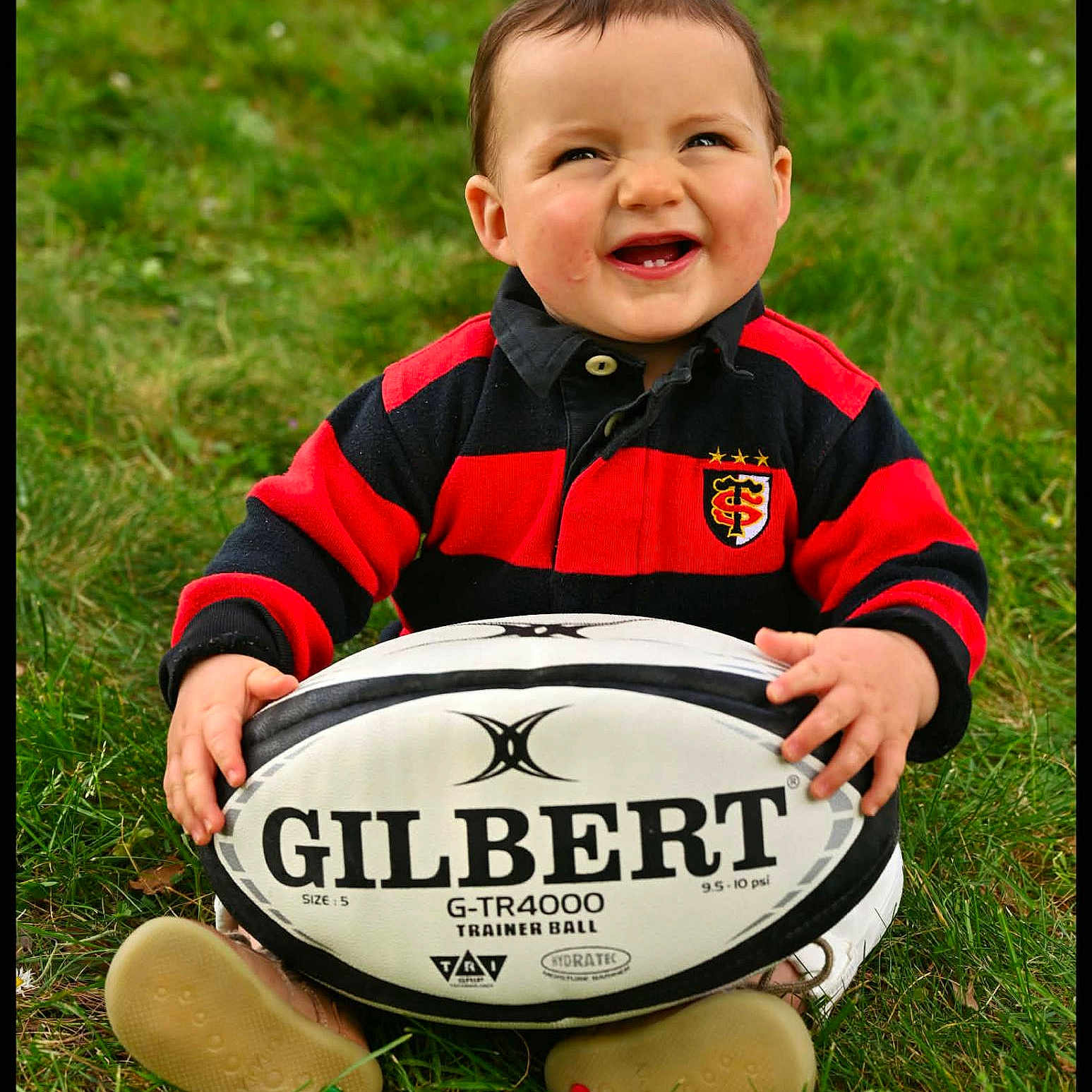 Jules participe au concours pour gagner de l'argent avec cette photo : toddler, child, grass, rugby_ball, smile, happy, outdoor, red_sweater, black_sweater, baby, playing, toy, sport, sitting, cute, portrait, person, fun, daylight, casual_clothing
