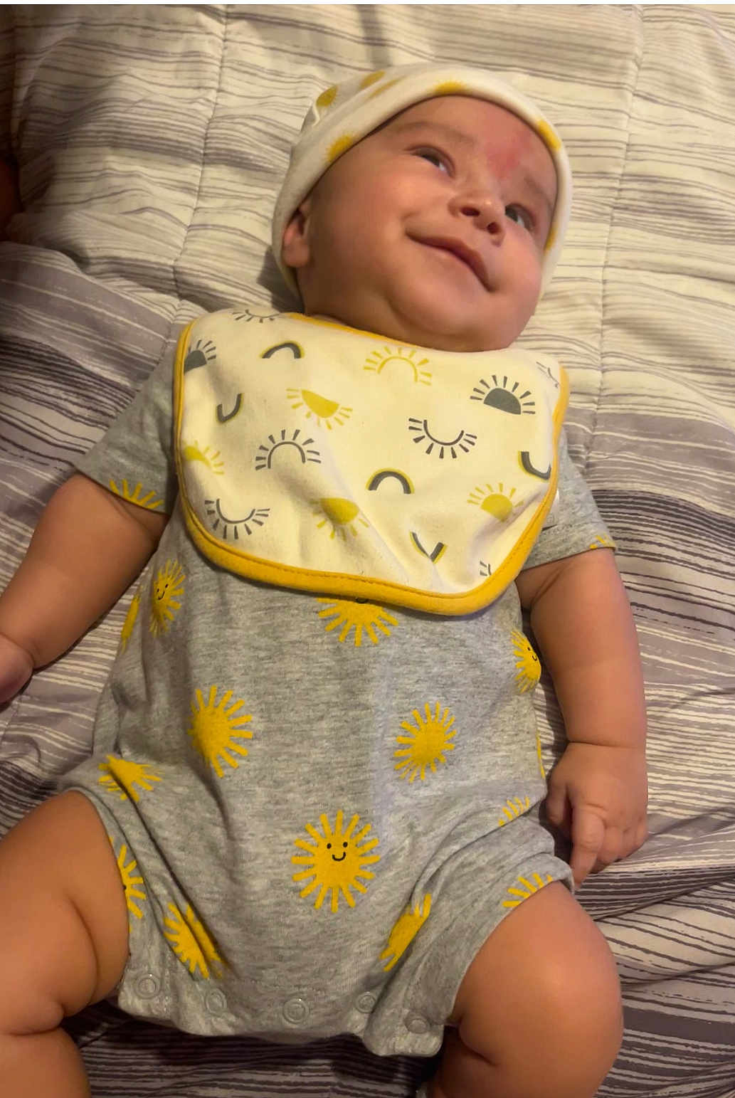 Calixto is registered to the contest to win money with this photo: baby, infant, smiling, onesie, hat, bed, bedding, striped, pattern, sun, yellow, grey, cute, child, lying_down, happy, face, skin, hand, chubby