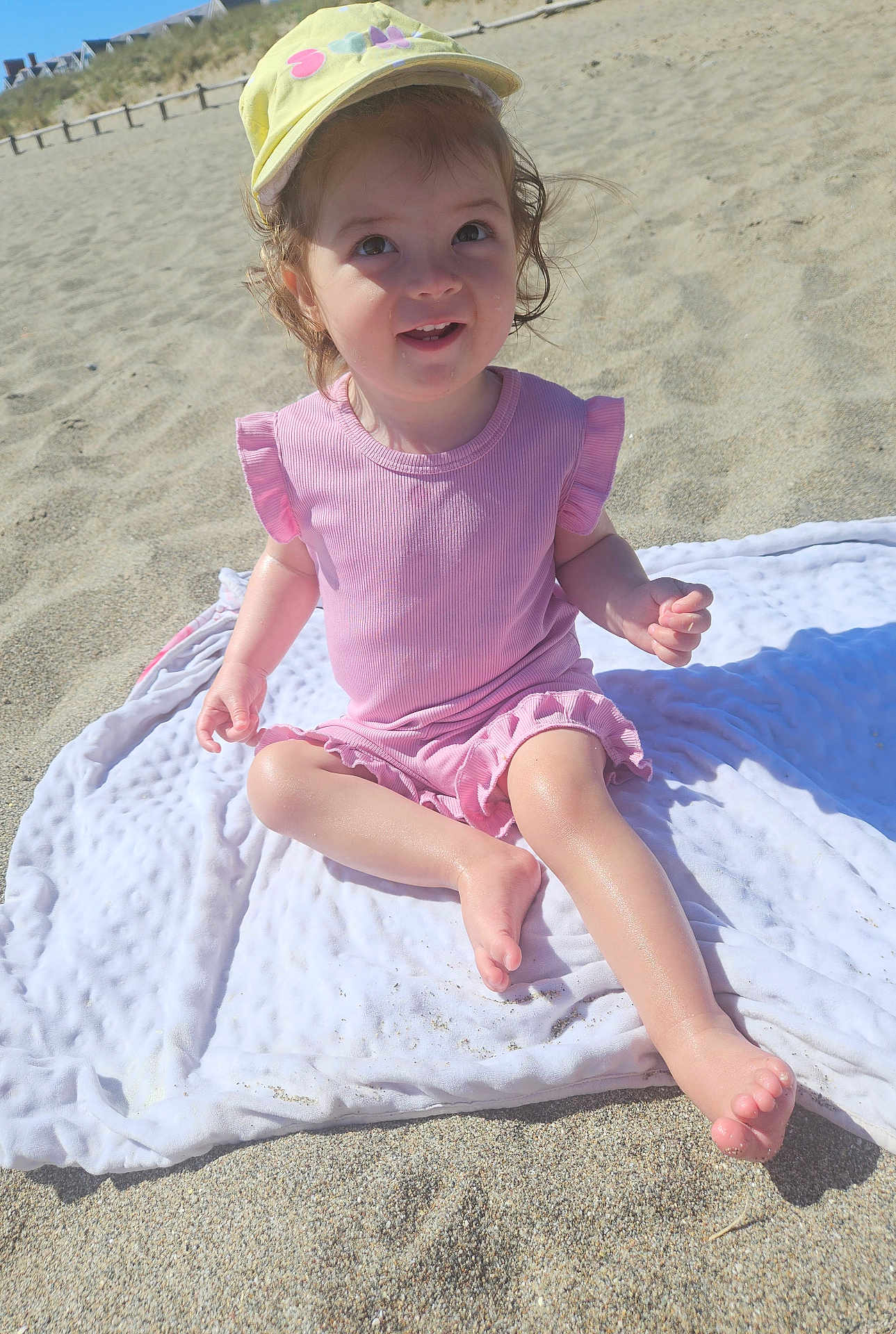 Ruby joined the competition — help win amazing prizes! child, toddler, beach, sand, blanket, hat, yellow_hat, pink_clothing, smiling, happy, sunlight, outdoor, summer, barefoot, cute, person, portrait, sitting, playful, daytime