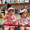 Josefina-Ravenna is registered to the contest to win money with this photo: toddler, child, shopping_cart, costco, warehouse, hat, pink_clothing, purple_clothing, water_bottle, curly_hair, blond_hair, blue_eyes, waving, sitting, retail_store, shelves, boxes, floor, casual, indoor