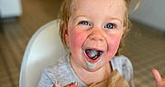 Josefina-Ravenna is registered to the contest to win money with this photo: toddler, child, ice_cream, messy, high_chair, napkin, happy, blue_eyes, face, hands, mouth_open, indoor, table, hair_tied, casual_clothing, smiling, food, dining, cute, playful