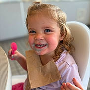 Josefina-Ravenna is registered to the contest to win money with this photo: toddler, child, high_chair, bib, smile, blue_eyes, curly_hair, messy_face, pink_spoon, indoor, happy, cute, food, napkin, portrait, person, clothing, table, hand, skin