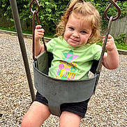 Josefina-Ravenna is registered to the contest to win money with this photo: child, toddler, swing, playground, outdoor, greenery, woodchips, smile, curly_hair, blonde_hair, blue_eyes, holding, metal_chain, green_shirt, black_shorts, happy, person, summer, fun, park