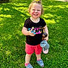 Josefina-Ravenna is registered to the contest to win money with this photo: toddler, child, grass, outdoor, smiling, happy, pacifier, toy, doll, watering_can, shoe, shorts, tshirt, hair_tie, sunny, daytime, greenery, playful, standing, casual_clothing