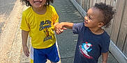 Rakim joined the competition — help win amazing prizes! child, children, holding_hands, smiling, sidewalk, outdoor, sunny, fence, curly_hair, yellow_shirt, blue_shorts, blue_shoes, toddler, happy, casual_clothing, person, white_shoes, graphic_tshirt, shadow, playful