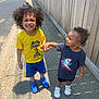 child, children, holding_hands, smiling, sidewalk, outdoor, sunny, fence, curly_hair, yellow_shirt, blue_shorts, blue_shoes, toddler, happy, casual_clothing, person, white_shoes, graphic_tshirt, shadow, playful