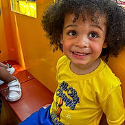Rakim is registered to the contest to win money with this photo: child, toddler, curly_hair, yellow_shirt, blue_shorts, smiling, indoor, play_area, bench, happy, person, cute, portrait, bright_colors, daylight, window, casual_clothing, seated, young_child, joyful