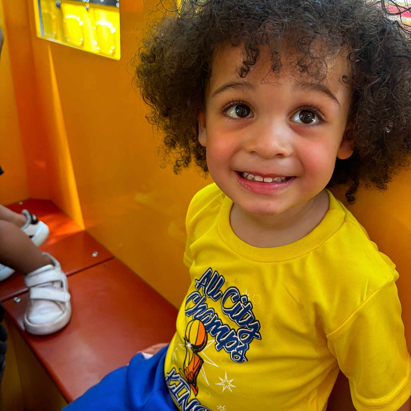 Rakim is registered to the contest to win money with this photo: bench, blue_shorts, bright_colors, casual_clothing, child, curly_hair, cute, daylight, happy, indoor, joyful, person, play_area, portrait, seated, smiling, toddler, window, yellow_shirt, young_child