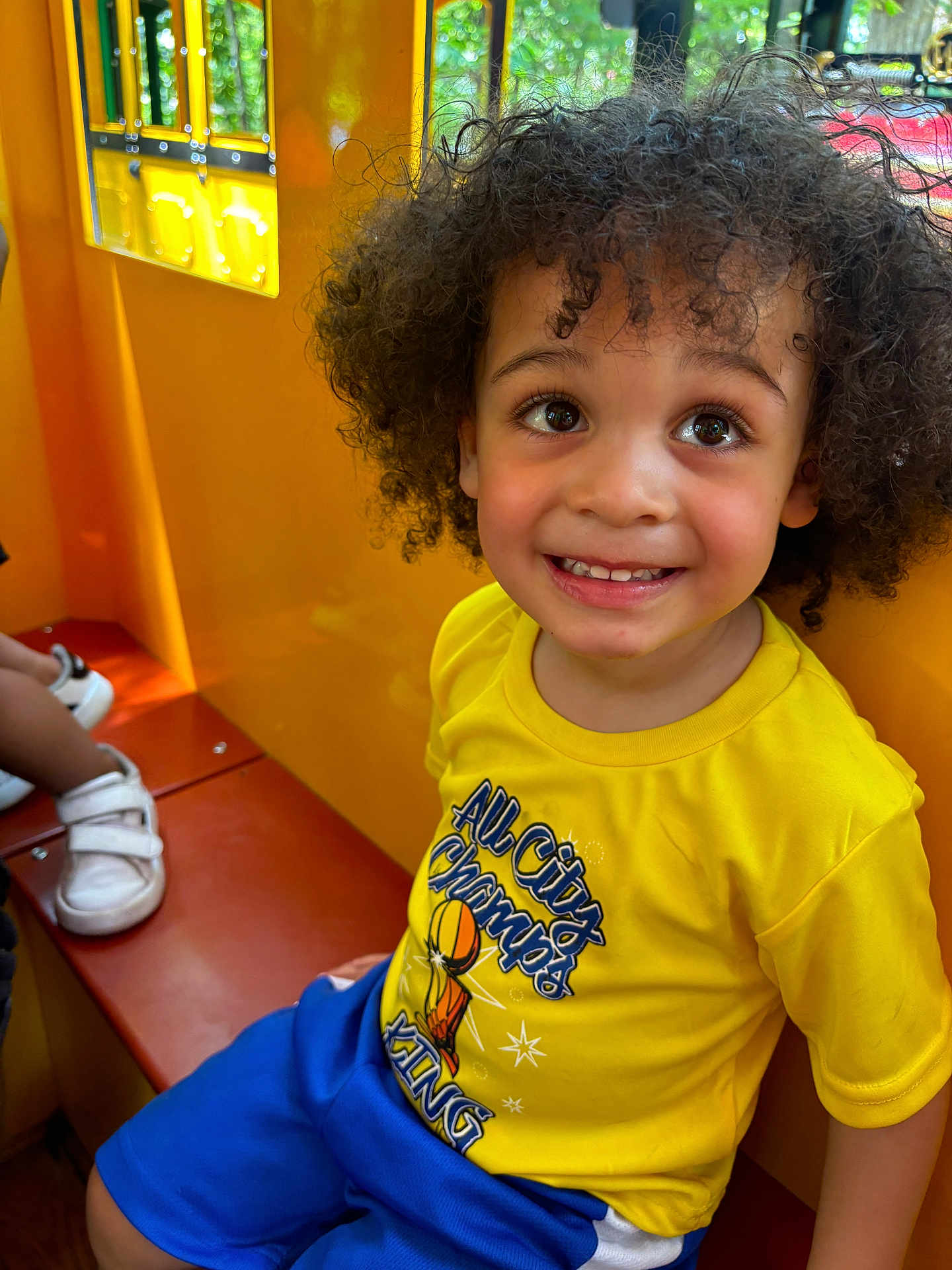 Rakim is registered to the contest to win money with this photo: child, toddler, curly_hair, yellow_shirt, blue_shorts, smiling, indoor, play_area, bench, happy, person, cute, portrait, bright_colors, daylight, window, casual_clothing, seated, young_child, joyful