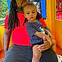 child, adult, man, toddler, dreadlocks, cap, red_shirt, blue_tshirt, playground, play_structure, yellow, blue, orange, outdoor, greenery, seated, holding, curious, casual_clothing, family