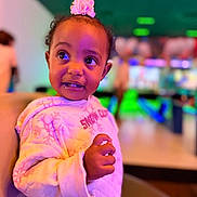 Sophie joined the competition — help win amazing prizes! baby, child, toddler, portrait, smiling, flower_headband, pink_clothing, indoor, colorful_lighting, neon_lighting, arcade, entertainment_venue, seated, hands, face, eyes, curly_hair, fashion, closeup, background_blur