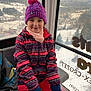 child, girl, winter_clothing, beanie, pompom_hat, jacket, mittens, snow, gondola, ski_resort, window, mountains, village, smiling, portrait, travel, cold_weather, seat, colorful_pattern, scenic_view